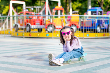 a child girl in an amusement park rides a skateboard in the summer and smiles with happiness near...