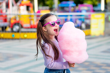 a child girl in an amusement park in the summer eats cotton candy near the carousels in sunglasses,...