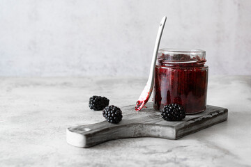 A glass jar with blackberry jam on a light wooden plank. Large blackberries on a marble table. A white spoon in the background. Light background, space for text © Анна Никитина