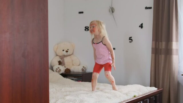 Little girl with painted face by red marker having fun on bed. Children room with bear and large clock in form numbers on wall.