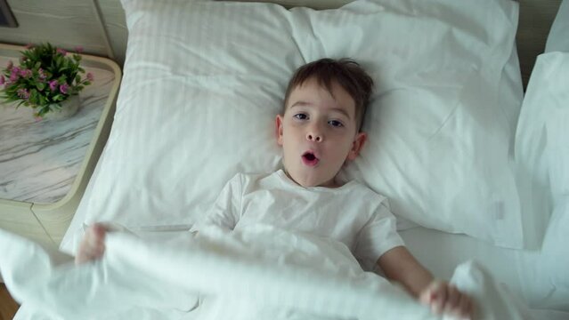 Portrait of a happy cute funny cheerful little child boy in white pajamas wakes up in bed in the morning, looks at his parents smiling, stretches laughs and hides under the blanket again.