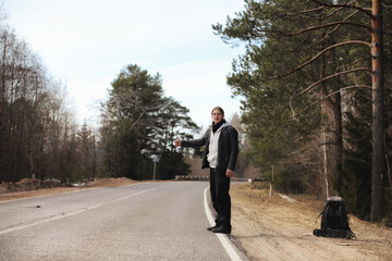 A young man is hitchhiking around the country. The man is trying to catch a passing car for traveling. The man with the backpack went hitchhiking to the south.