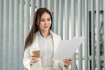 Asian businesswoman analyzing documents (data graph) and sipping coffee while working in a private office, Happy working lifestyle, Modern private office