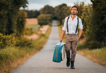 Individual man in vintage clothing holding a suitecase is walking along a street in nature