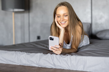 Fun young woman lying on her stomach hold in hand use phone relax in bed