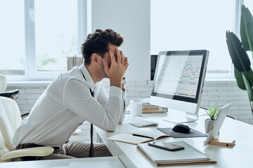 Depressed young man covering face with hands while sitting at his working place in office