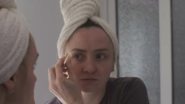 Girl removes excess hair on eyebrows with tweezers in front of mirror. Young woman with towel on head in bathroom near the mirror. Morning routine