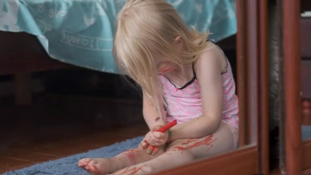 Little girl with blonde hair sits on floor and enthusiastically paints arms and legs with red felt-tip pen.