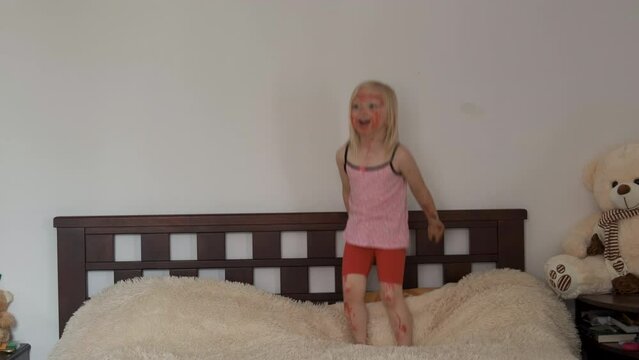 Little girl fidget all painted with red marker. Cheerful kid jumps high on soft bed. Happy childhood.