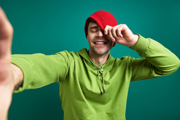 Fototapeta premium Happy young man making selfie and pulling hat on face while standing against green background