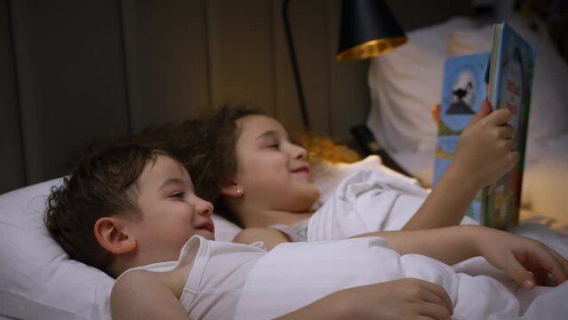 Child care at home, little sister takes care of younger brother, reads a bedtime story to children, Leisure time for children. Daughter reading a book to her brother while lying in bed at home