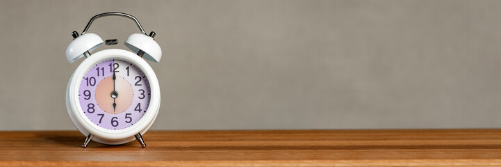 Six o'clock on the alarm. A white alarm clock stands on a wooden table. The clock hand points to six o'clock. Switching to summer or winter time. Set an alarm for 6:00 or 18:00. 