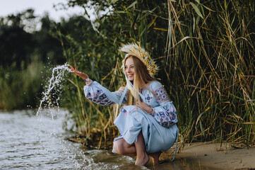 A beautiful, young happy, cheerful blonde Ukrainian girl in an embroidered, long dress with a wreath of wheat and wild flowers sits near the sea and splashes water. Photography, Ukraine.