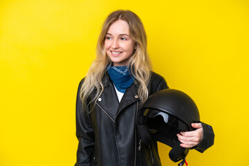 Blonde English young girl with a motorcycle helmet isolated on yellow background looking to the side and smiling