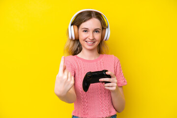 Blonde English young girl playing with a video game controller isolated on yellow background doing coming gesture