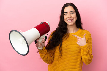 Young caucasian woman isolated on pink background holding a megaphone and pointing side