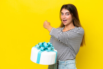 Young caucasian woman holding birthday cake isolated on yellow background pointing back