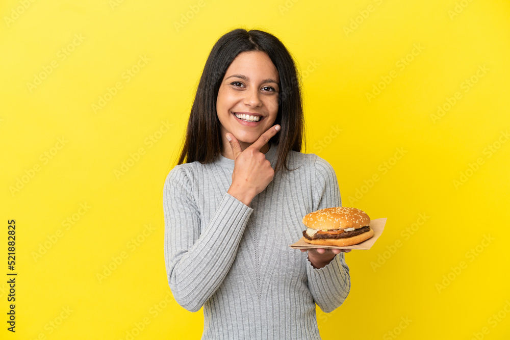 Wall mural Young caucasian woman holding a burger isolated on yellow background happy and smiling