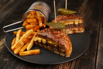 Delicious beef burger cut in half with cheese, pickled, tomatoes, mustard, and fries on a black plate on wooden background. Fast food.