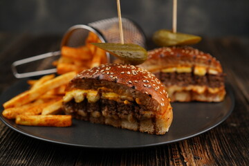 Delicious beef burger cut in half with cheese, pickled, tomatoes, mustard, and fries on a black plate on wooden background. Fast food.