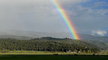 Bright rainbow over pine-covered hills and grassland, overcast day with distant mountains under grey clouds, in Montana, USA - Powered by Adobe