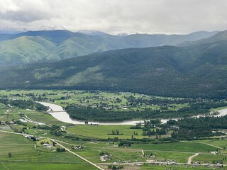 Fototapeta na wymiar Aerial view of the Bitterroot River flowing through lush, green fields, next to homes, hills and mountains in Missoula area, Montana, USA. Rain clouds overhead