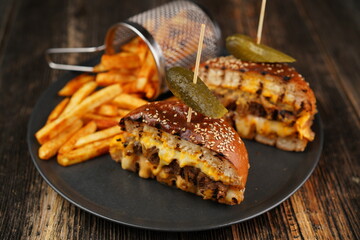 Delicious beef burger cut in half with cheese, pickled, tomatoes, mustard, and fries on a black plate on wooden background. Fast food.