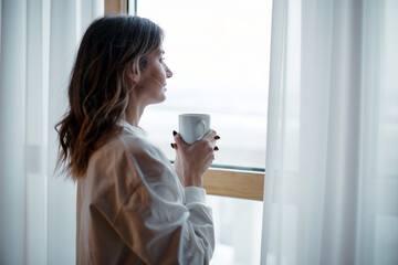 Romantic dreamy female homeowner tenant holding mug of hot coffee looking out of window. Leisurely morning