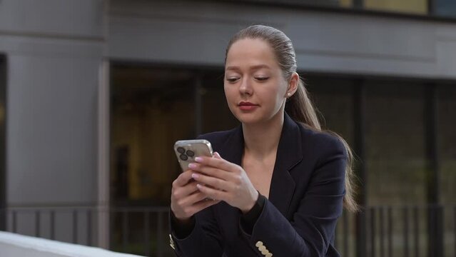A female realtor is holding her phone while leaning on the parapet in a modern urban space in the financial district. A businesswoman in the contemporary garden in a cluster of tall buildings.