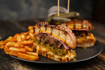 Delicious beef burger cut in half with cheese, pickled, tomatoes, mustard, and fries on a black plate on wooden background. Fast food.