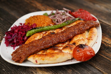 Traditional Turkish Adana kebab. Adana kebab with onions, bulgur, roasted green pepper, and roasted tomato on a white plate on a wooden table. Turkish Adana kebab with special restaurant presentation.