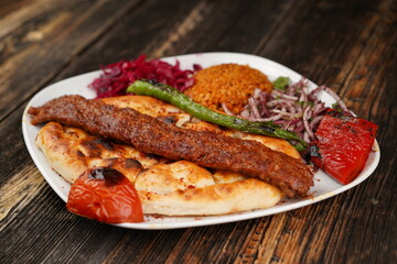 Traditional Turkish Adana kebab. Adana kebab with onions, bulgur, roasted green pepper, and roasted tomato on a white plate on a wooden table. Turkish Adana kebab with special restaurant presentation.