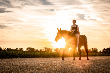 Junge Frau reitet im Sonnenuntergang über das Stoppelfeld