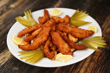 Crispy deep-fried chicken strips. Fried chicken strips on fried potatoes with pickles. Fried chicken and potatoes on a white plate on wooden background. 