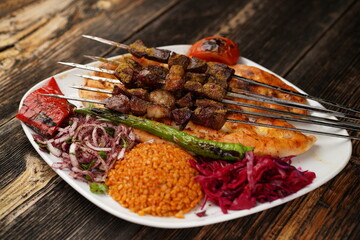 A skewered meat kebab with grilled tomato, onion, and rise. Grilled shish meat served with a white plate on wooden background. Traditional grilled kebabs.