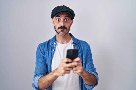 Hispanic man with beard using smartphone typing message puffing cheeks with funny face. mouth inflated with air, catching air.