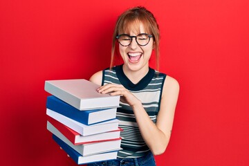 Redhead young woman holding a pile of books smiling and laughing hard out loud because funny crazy joke.