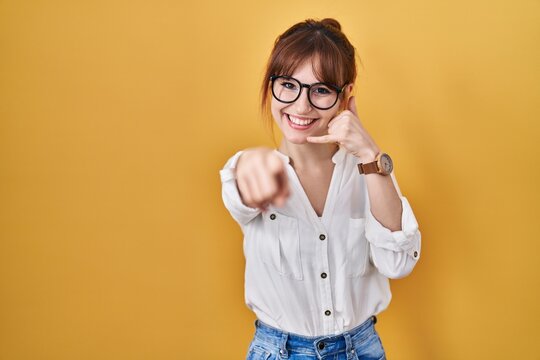 Young beautiful woman wearing casual shirt over yellow background smiling doing talking on the telephone gesture and pointing to you. call me.