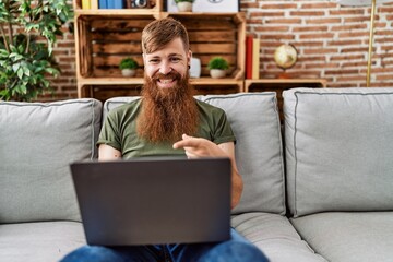 Redhead man with long beard using laptop sitting on the sofa at the living room smiling happy pointing with hand and finger