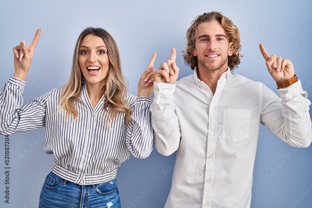 Poster young couple standing over blue background smiling amazed and surprised and pointing up with fingers