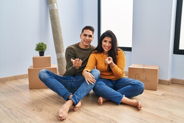 Young couple sitting on the floor at new home beckoning come here gesture with hand inviting welcoming happy and smiling