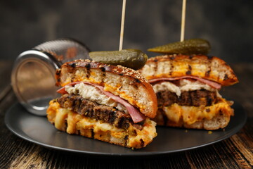 Delicious beef burger cut in half with cheese, pickled, tomatoes, mustard, and fries on a black plate on wooden background. Fast food.  