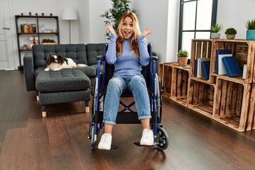 Fototapeta premium Young beautiful woman sitting on wheelchair at home celebrating crazy and amazed for success with arms raised and open eyes screaming excited. winner concept