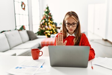 Young caucasian girl sitting on the table working using laptop by christmas tree doing stop sing with palm of the hand. warning expression with negative and serious gesture on the face.