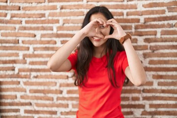 Young teenager girl standing over bricks wall doing heart shape with hand and fingers smiling looking through sign