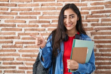 Young teenager girl wearing student backpack and holding books smiling happy pointing with hand and finger to the side