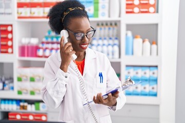 African american woman pharmacist talking on telephone reading document at pharmacy - Powered by Adobe
