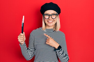 Beautiful blonde woman wearing french look with beret holding painter brushes smiling happy pointing with hand and finger