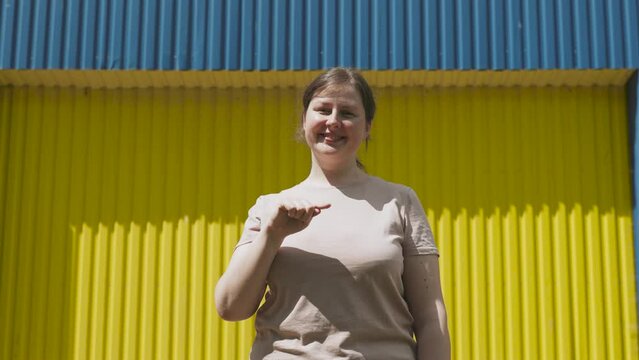 Simple slightly disheveled caucasian woman draws heart shape with fingers in air. Lady shows with her hands symbol of heart, peace and love against bright blue and yellow wall on sunny day.