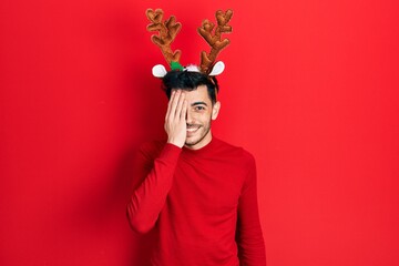 Young hispanic man wearing cute christmas reindeer horns covering one eye with hand, confident smile on face and surprise emotion.